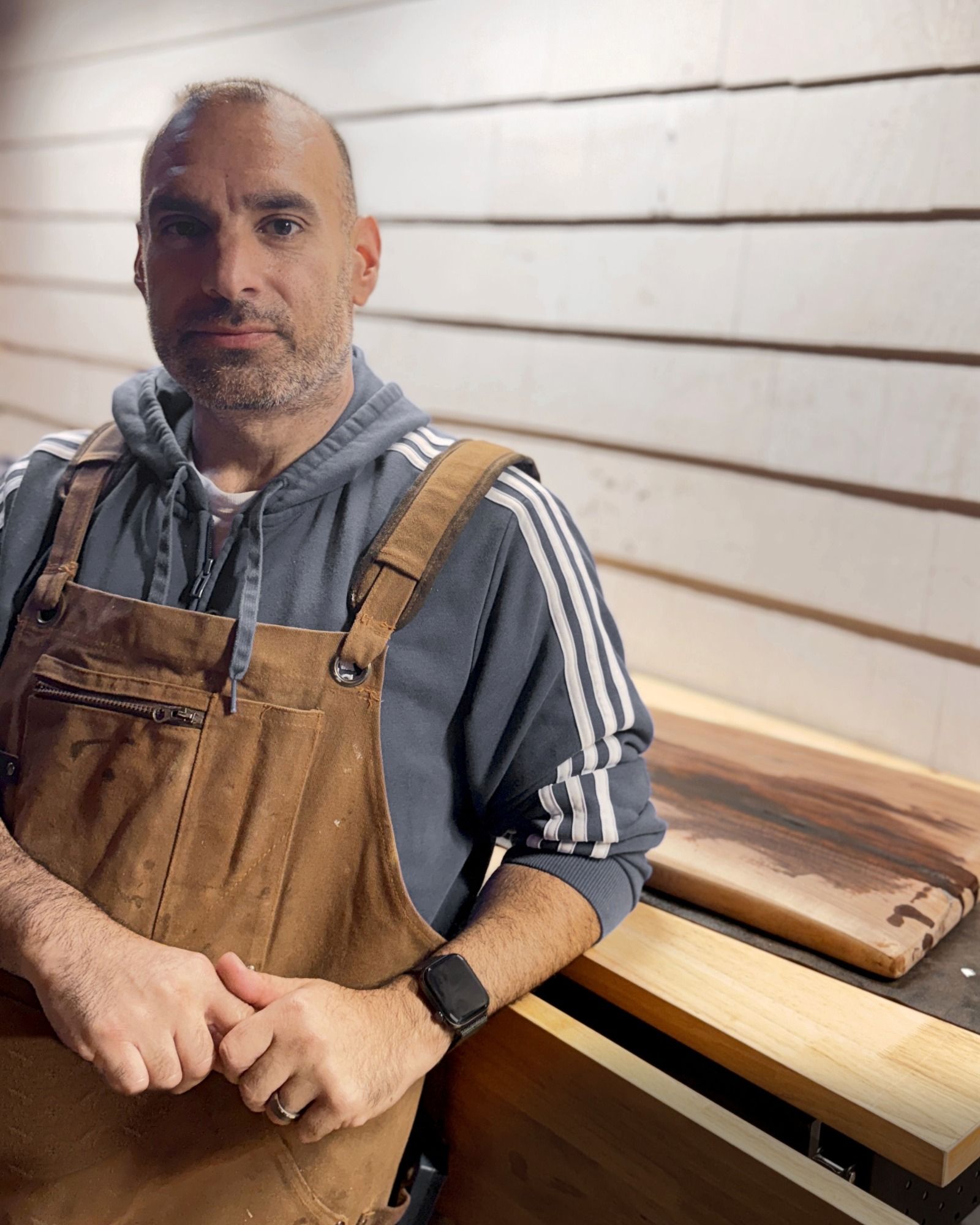 Jim Tomasso, founder of Black Walnut Co., leaning on a workbench in his shop wearing a canvas apron, with a finished walnut piece behind him.